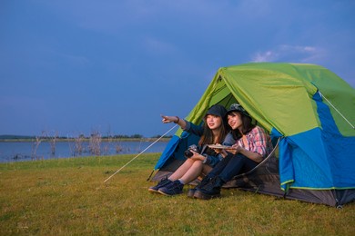 group of asian friends tourist drinking and playing guitar together with happiness in summer while having camping near lake at sunset