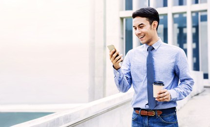 smiling young asian businessman using mobile phone in the city. modern urban lifestyle. male portrait. hand holding coffee cup. looking at smartphone