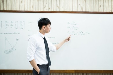 an asian male teacher in a classroom near the whiteboard is writing and teaching a lesson with a special lecture in a class column. math teacher explains arithmetic sums to university students.