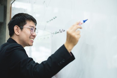an asian male teacher in a classroom near the whiteboard is writing and teaching a lesson with a special lecture in a class column. math teacher explains arithmetic sums to university students.