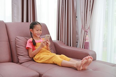 happy asian little girl child while playing smartphone on sofa in living room at home