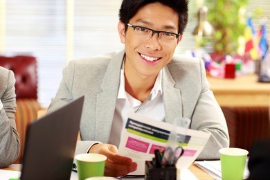 happy asian businessman in glasses sitting at the table and holding document in office