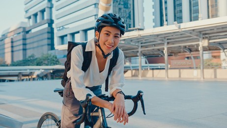 asian businesswoman go to work at office stand and smiling wear backpack look at camera with bicycle on street around building on a city. bike commuting, commute on bike, business commuter concept.
