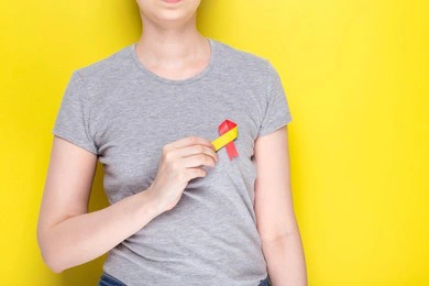 world hepatitis day concept. girl in gray t-shirt holds in her hand awareness symbol red-yellow ribbon. yellow background. copy space