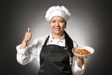 beautiful asian chef with dish showing thumb-up gesture on dark background