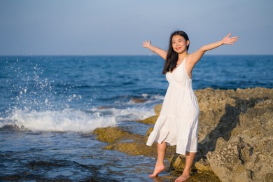 summer lifestyle portrait of young beautiful and happy asian woman at the beach - attractive korean girl by the sea enjoying holidays trip on rock cliff with ocean view