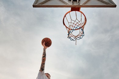 hand of man throwing basketball to the basket hoop. bottom up view of basketball hoop and ball against cloudy sky