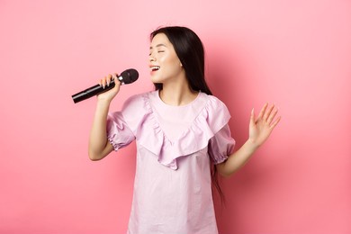 beautiful asian girl perform song, singing in microphone and smiling romantic, standing in dress against pink background