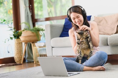 happy cheerful asain woman wear headset laughing while holding her cat during using laptop for video conference in living room . working at home concept
