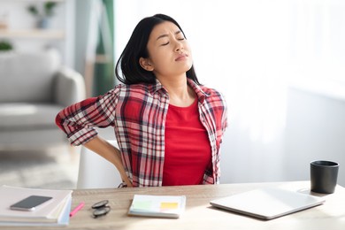 beautiful young asian woman suffering from backache while sitting at desk in home office, tired korean freelancer lady having acute lower back pain after long time working with laptop computer
