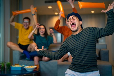group of asian people friends sit on sofa watching and cheering sports games competition on tv together at home. excited man and woman sport fans celebrate sport team victory in national sports match