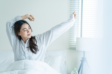 asian beautiful girl in pajamas wake up in the morning with happiness. attractive young woman smiling, feel happy and relax then stretching body after getting up from sleep on bed in bedroom at home.