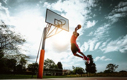 street basketball player making a powerful slam dunk on the court - athletic male training outdoor at sunset - sport and competition concept
