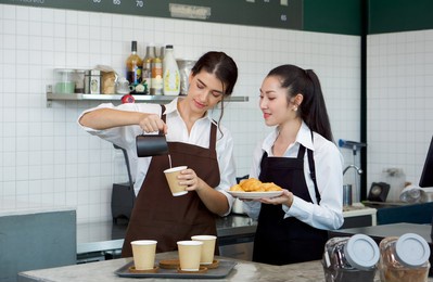 young caucasian barista pouring milk from the jug into a paper coffee cup. asian assistant in an apron holding croissant plate stands beside her. morning atmosphere in a coffee shop.