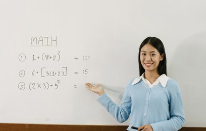 asian female teacher is teaching students at the classroom while pointing at numbers on the white board.