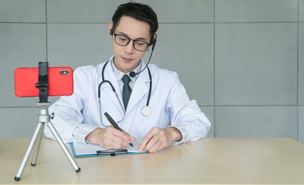 male doctor consult patient with video call on smartphones,  wearing earphones and taking notes health information. asian man physician therapist talking online telemedicine medical consultation.