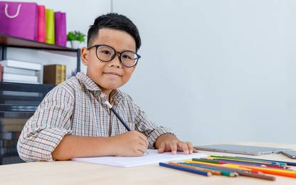 smart young asian boy wearing glasses and grey checked shirt sitting on studying desk of tablet, mobile phone, and colorful pencils in reading room and concentrate on drawing on white paper