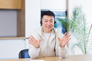 asian man talking online on video call at home room with headset. male in casual clothes looking at camera, meeting or conference in distance. portrait student or teacher lecture training. webcam view