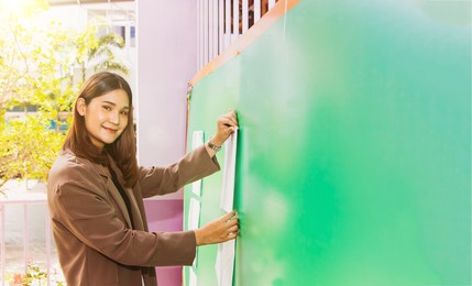 shot portrait asian female teacher stands and prepares a bulletin board to post an announcement on a blank green board for students to read in middle school.