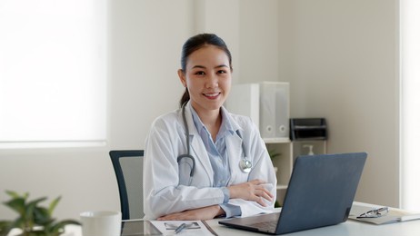 young adult asia people or medic staff woman sitting toothy smile arm cross look at camera happy work on desk in clinic office room in telehealth telemedicine smart health care consult online service.