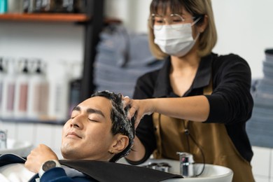 asian attractive young male get hair washed by hairdresser in salon. expert hair stylist woman wear mask enjoy massage handsome man customer who's lying down on salon washing bed at beauty barber shop