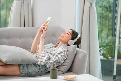 photo of modern asian woman wearing headphones holding cell phone while lying on sofa in bright apartment