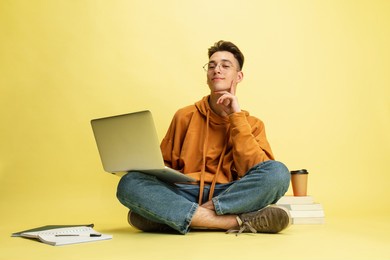 studying, doing homework. one young smiling caucasian man, student in glasses sits on floor with laptop isolated on yellow studio background. education, studying and student life concept.