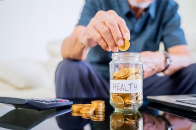 close up of senior man hands savings coins in a glass jar with health word on the table
