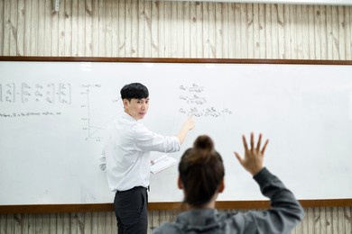 an asian male teacher in a classroom near the whiteboard is writing and teaching a lesson with a special lecture in a class column. math teacher explains arithmetic sums to university students.