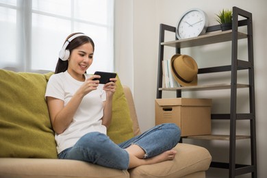 beautiful young asian women play mobile game and put on wireless headphone sitting on sofa in living room. playing game on smartphone winning victory moment. very enjoy and fun relax time
