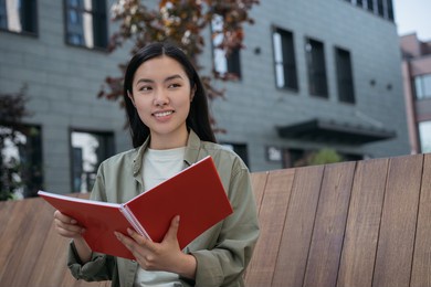 portrait of pensive asian business woman planning start up project sitting on bench looking away. smiling student studying, holding book, exam preparation in university campus. education concept	