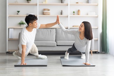 joyful millennial asian man and woman in sportswear cheering up each other while planking at home, giving high five and smiling, doing exercises on fitness mats at living room, healthy lifestyle