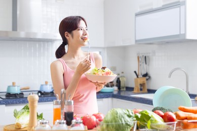 eat healthy - smiling asian pretty female eating salad with fruits and vegetables in kitchen