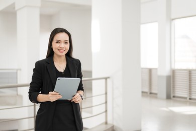 portrait stock photo of a confident cheerful asian businesswoman professional in black business suit using a digital tablet standing in the office building representing business oriented concept
