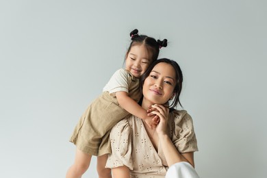 cheerful asian toddler daughter hugging happy mother isolated on gray