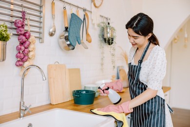 asian woman in protective gloves is wiping dust using a spray and a duster while cleaning her house. beautiful young female makes cleaning the kitchen. women doing housework, housewife portrait.
