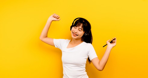 music and relaxation concept beautiful asian woman in headphones listening to music and dancing on yellow background happily in life