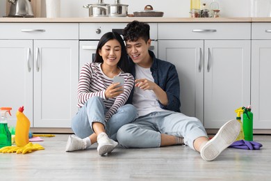 tired young asian man and woman sitting on floor among cleaning tools and supplies, using mobile phone and smiling, looking for house-keeping service online, on internet, copy space