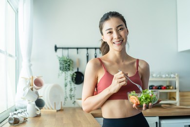 portrait of asian attractive woman hold salad bowl and look at camera. beautiful sport girl in sportswear enjoy eat clean vegetables after exercise for health in house. diet and healthy food concept.