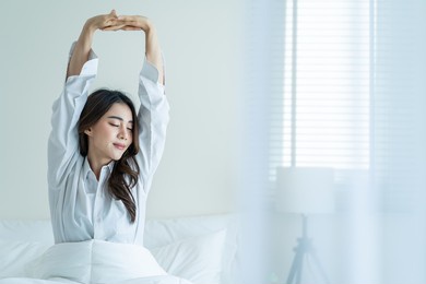 asian beautiful girl in pajamas wake up in the morning with happiness. attractive young woman smiling, feel happy and relax then stretching body after getting up from sleep on bed in bedroom at home.
