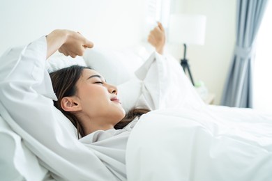 asian beautiful girl in pajamas wake up in the morning with happiness. attractive young woman smiling, feel happy and relax then stretching body after getting up from sleep on bed in bedroom at home.