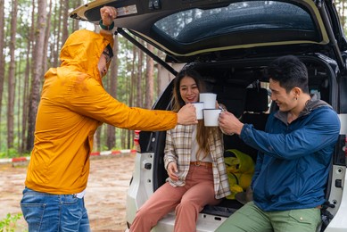 group of asian people friends sit on open car trunk talking and drinking coffee together at natural park. man and woman friendship enjoy outdoor lifestyle road trip and camping on summer vacation.