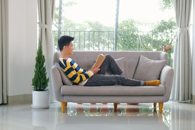 portrait of joyful young man reading book while sitting on sofa in his living room