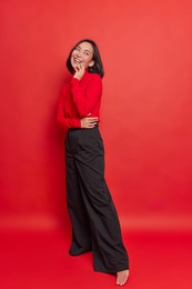 full length shot of happy dreamy brunette young asian woman has positive expression wears turtleneck black loose trousers stands against vivid red background thinks about something very pleasant
