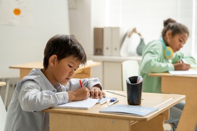 asian schoolboy making notes in copybook by desk against his classmate