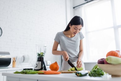 young asian woman cutting zucchini near raw vegetables and electric shaker in kitchen
