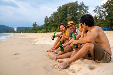 group of happy asian man and woman friends sitting on the beach enjoy drinking beer with talking together at summer sunset. male and female friendship relax and having fun outdoor lifestyle activity.