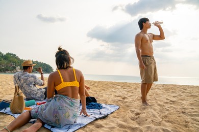 group of happy asian man and woman friends sitting on the beach enjoy picnic and talking together at summer sunset. male and female friendship relax and having fun summer outdoor lifestyle activity.