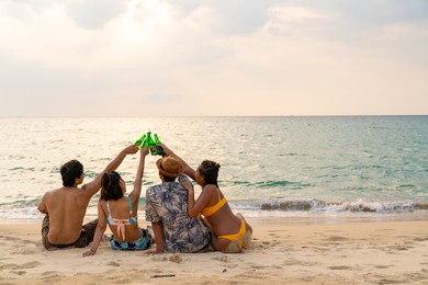 group of happy asian man and woman friends sitting on the beach enjoy drinking beer with talking together at summer sunset. male and female friendship relax and having fun outdoor lifestyle activity.