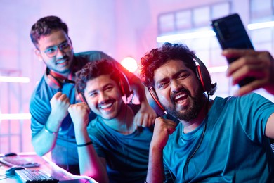 team of happy cheerful professional gamers with headphones vlogging, live streaming, or taking selfie on mobile phone in front of monitors during esports video gaming tournament